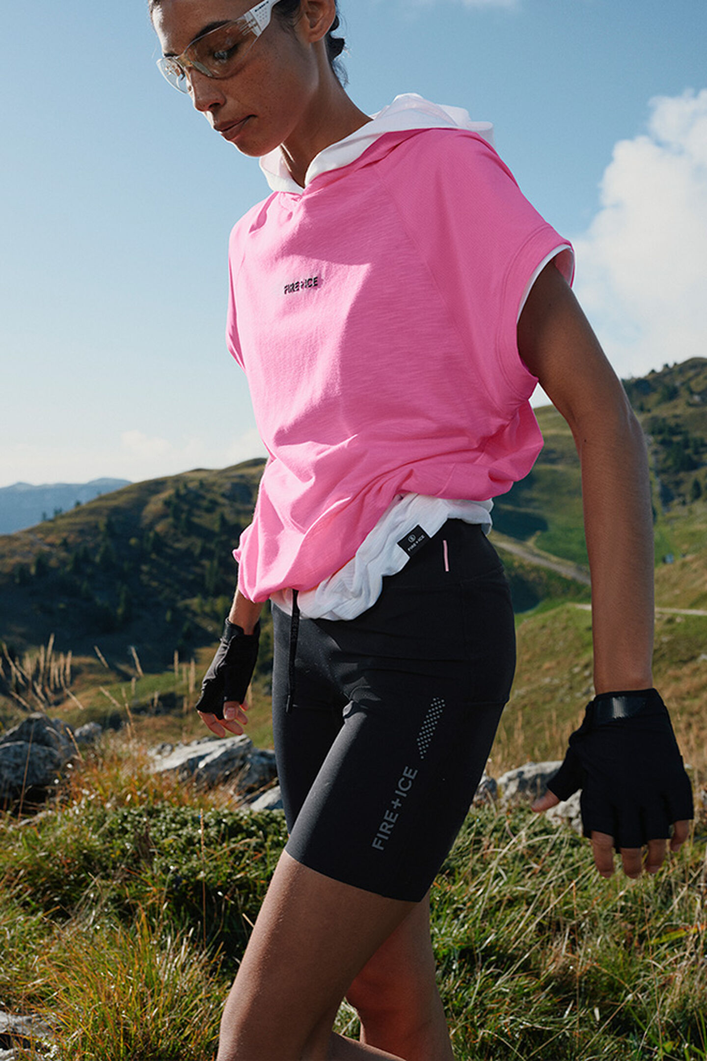The female model is hiking through the meadows of a mountain landscape. She is wearing pink short-sleeve sweatwear and cycling shorts.