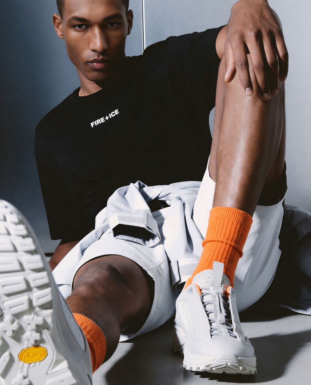Another male model is sitting on the ground. He is wearing white trainers that coordinate with his black and white outfit.