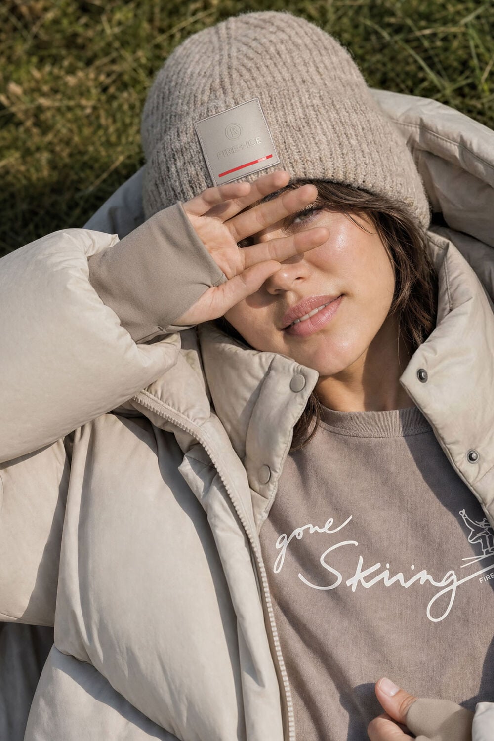 Das Damen Model sch&uuml;tzt ihr Gesicht vor der Sonne. Sie tr&auml;gt T-Shirt Kate mit &bdquo;Gone Skiing&ldquo;-Print, dazu eine M&uuml;tze und Daunenmantel Fleury mit Kapuze.