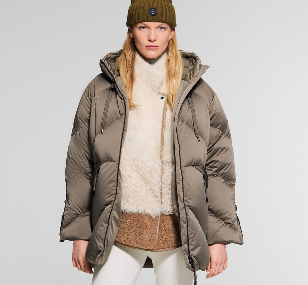 The blonde female model is wearing the Glenda down parka in taupe, over a teddy fur jacket and stirrup trousers. Green trousers complete the ensemble.