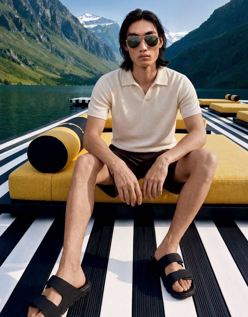 Das Herrenmodell sitzt auf einem in einem Bergsee liegenden Steg und tr&auml;gt das Strick-Polo-Shirt Finn in Off-White zu schwarzen Shorts und Sandalen.
