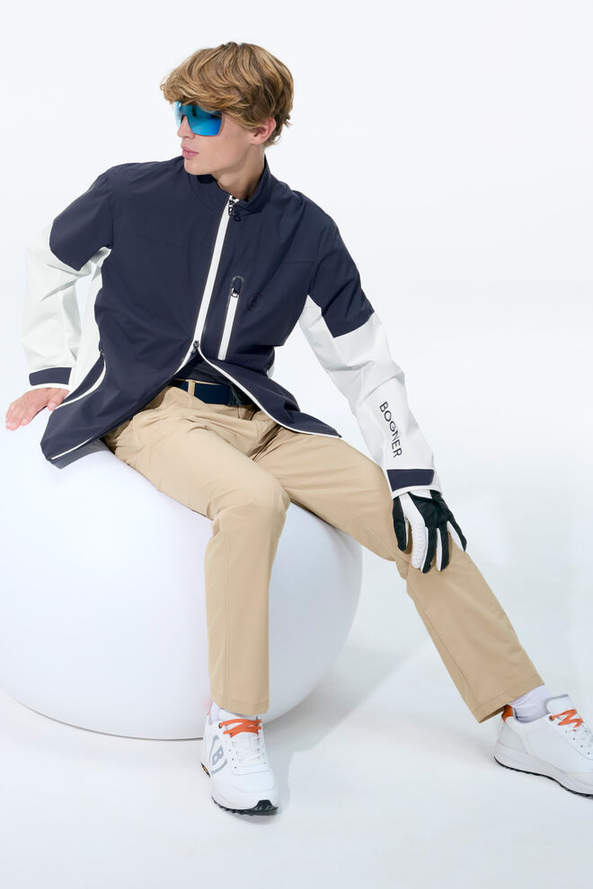 The seated male model is wearing a navy blue and white functional jacket, beige-coloured trousers and golf shoes.