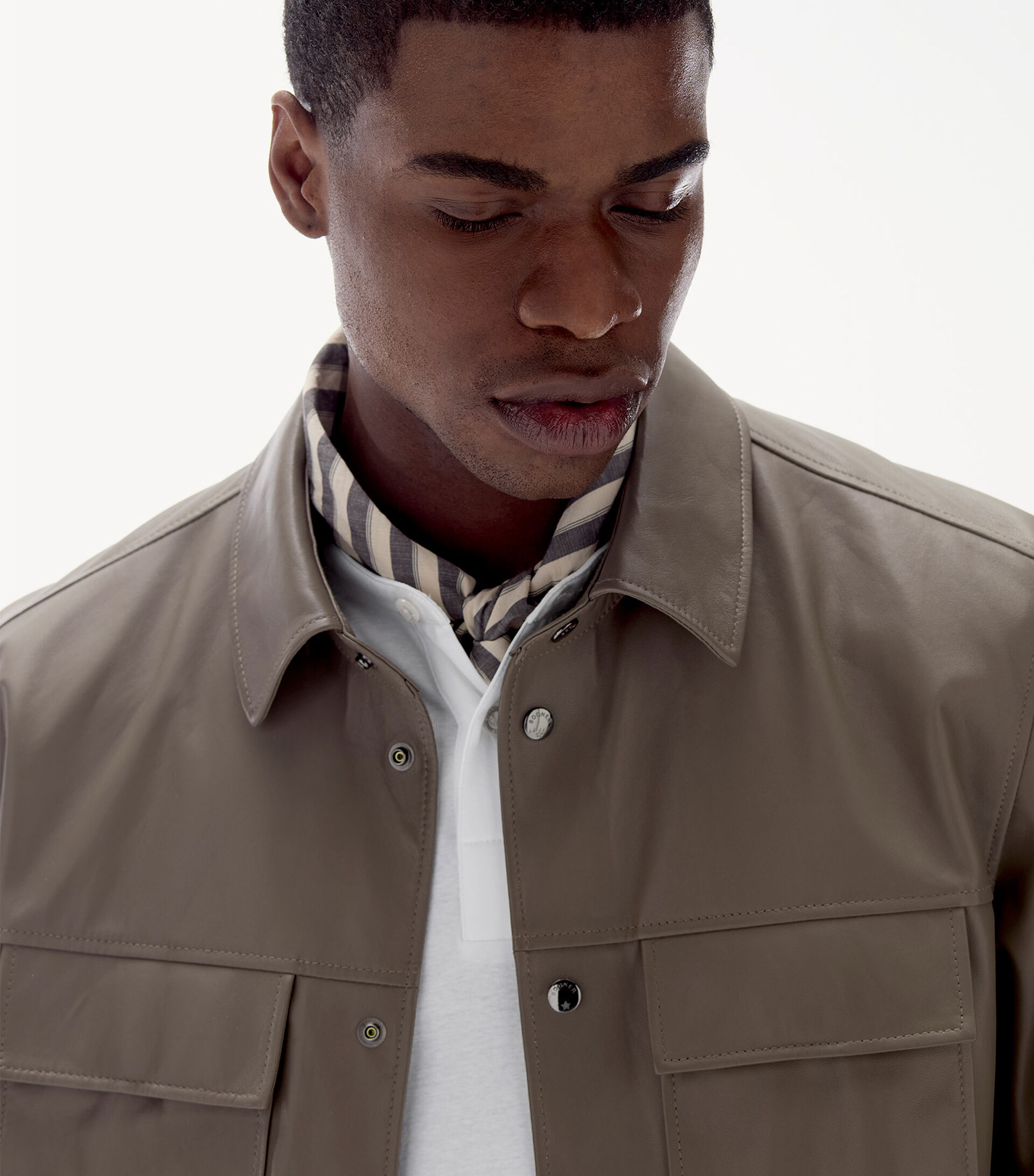 Close-up vom Herren Model. Er trägt ein Leder-Overshirt in Mud, darunter ein gestreiftes sowie ein reinweißes Hemd.