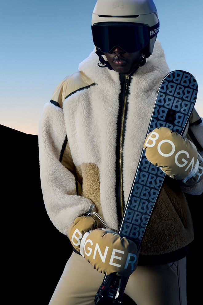 The model is wearing a luxurious ski jacket in beige teddy fur and is holding a ski in one hand. 