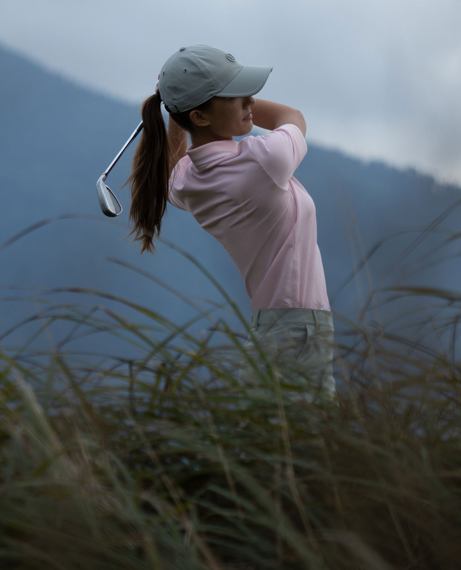 The female golf player is seen from the side. She is teeing off and wears a light pink polo shirt, a base cap and green pants.