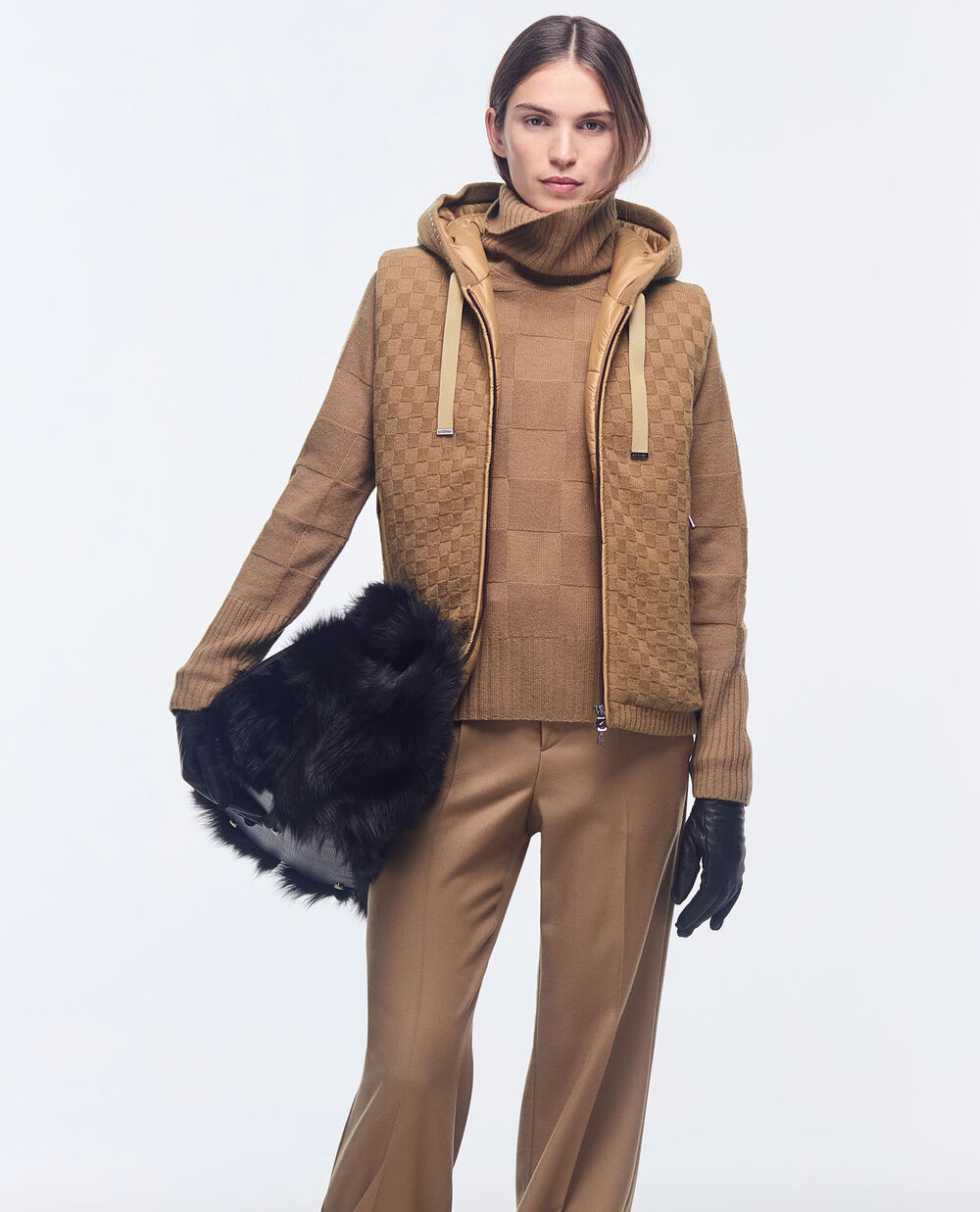The model is wearing a turtleneck pullover in camel with rectangular quilting, finished off with a knitted gilet and pleat-front trousers.