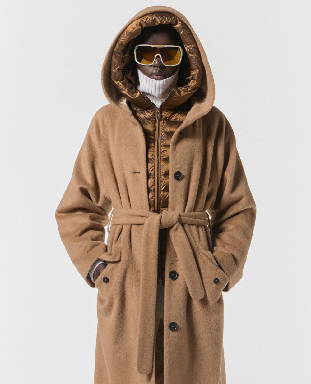 The model is wearing a down jacket with the hood over her head, with a camel-coloured coat, also with a hood, over the top.