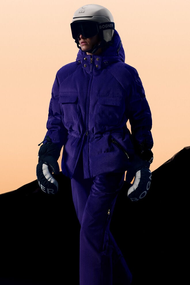 The model is standing in front of a mountain and wearing a purple cord ski outfit with black gloves. 