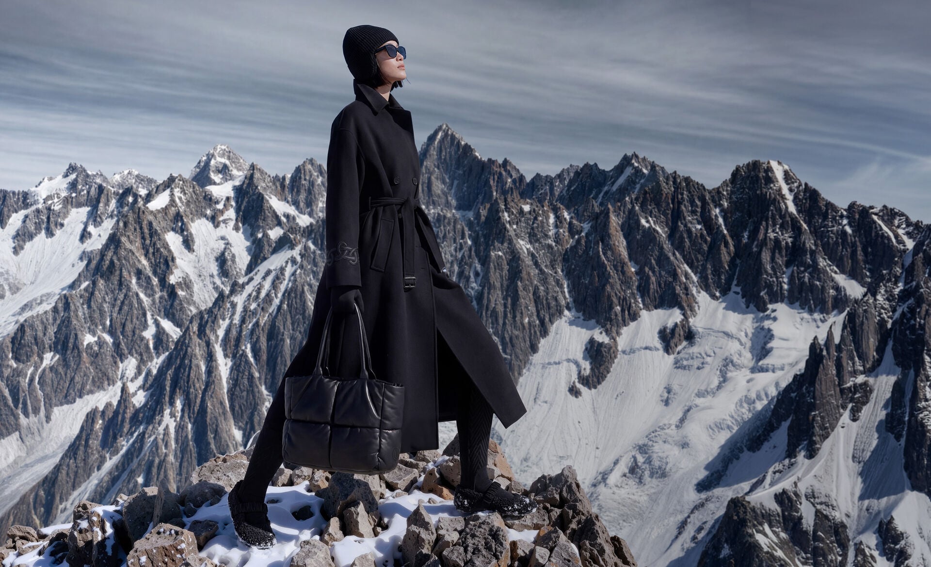 The female model with mountain scenery in the background is wearing a black coat with a wrap belt, and a knitted hat, sunglasses, a tote bag and teddy-fur ballerina flats to complete the look.