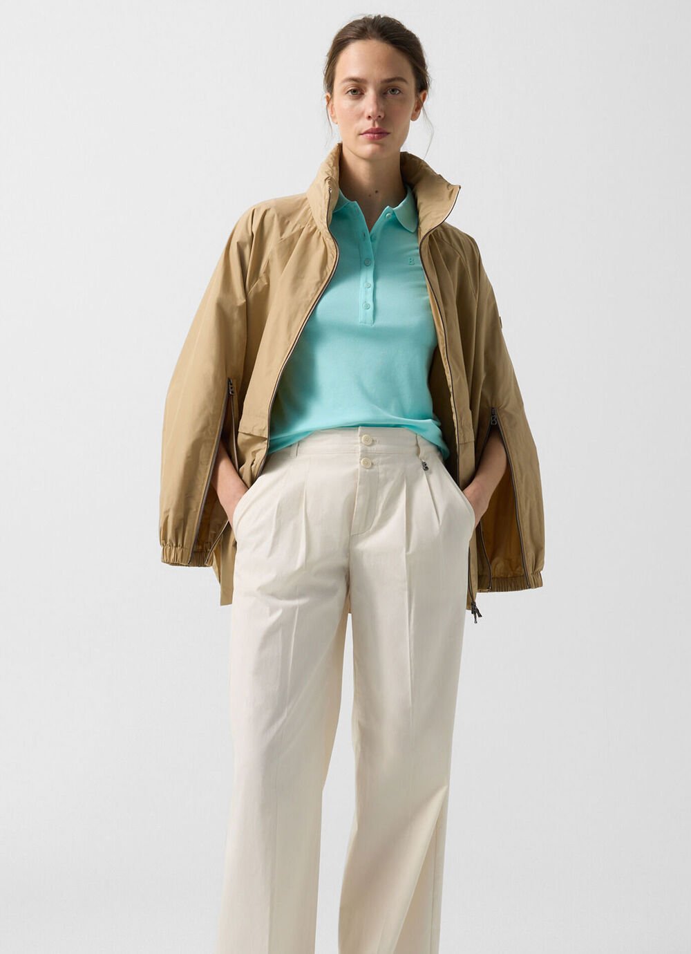 The Noelle jacket in camel is paired with a polo shirt in cyan blue and light-coloured trousers on the female model.