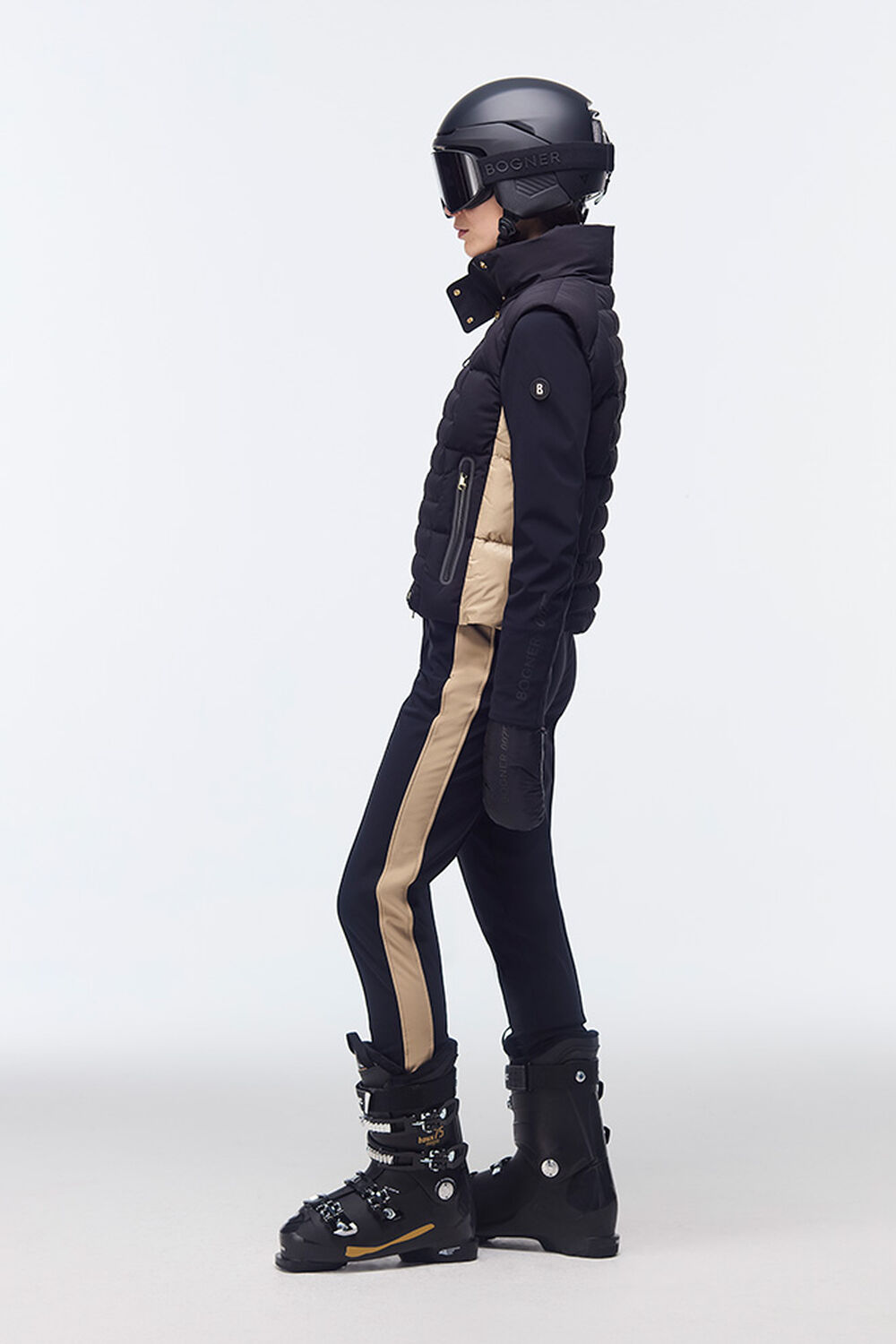 The model is standing sideways and wearing a ski look in Black/Beige with a black ski helmet, ski goggles and ski boots.