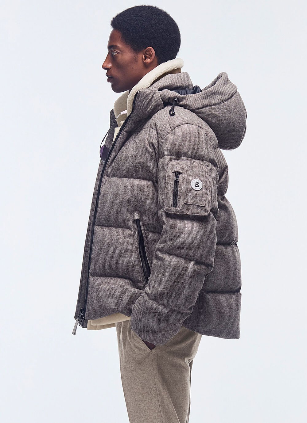 Het model draagt een grijze wollen donsjas met capuchon en een taupekleurige wollen broek.