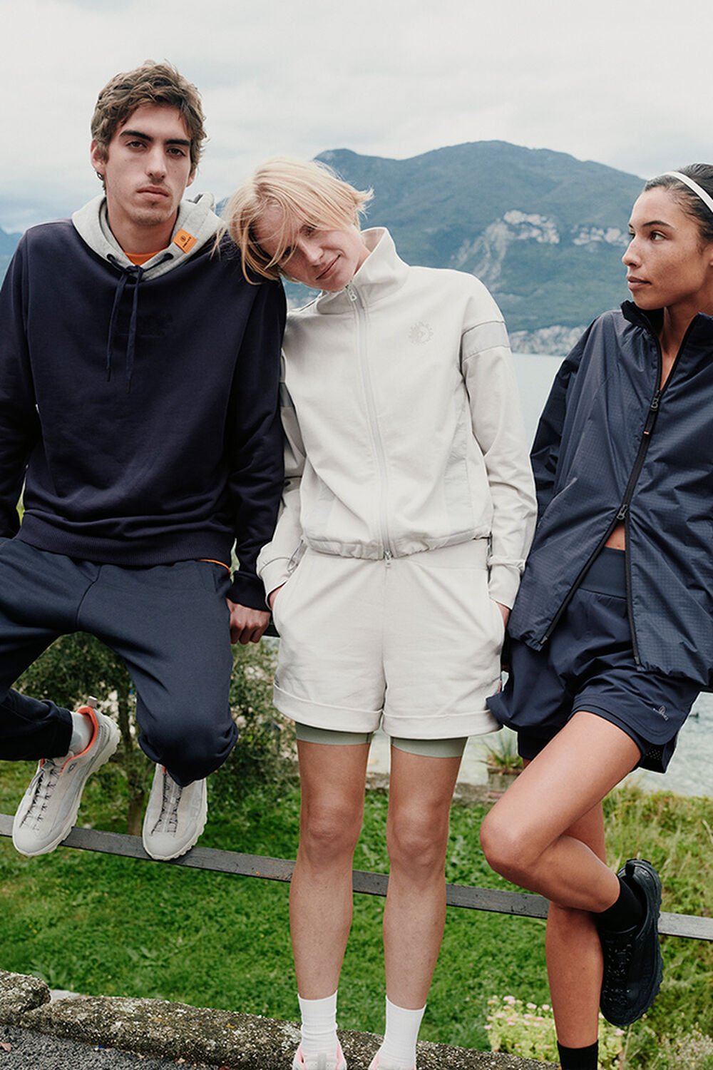 Eine Gruppe von drei Models in FIRE+ICE Looks in Navy und Beige. Sie lehnen vor einer Bergkulisse an einem Gel&auml;nder.