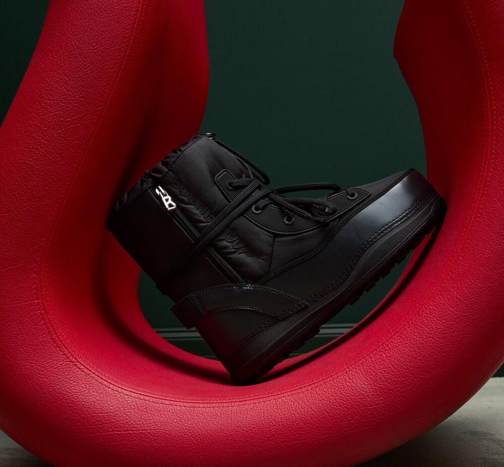 La chaussure d'hiver, posée en diagonale sur un objet déco rouge, est dotée d'une fermeture éclair argentée en forme de B et de lacets.