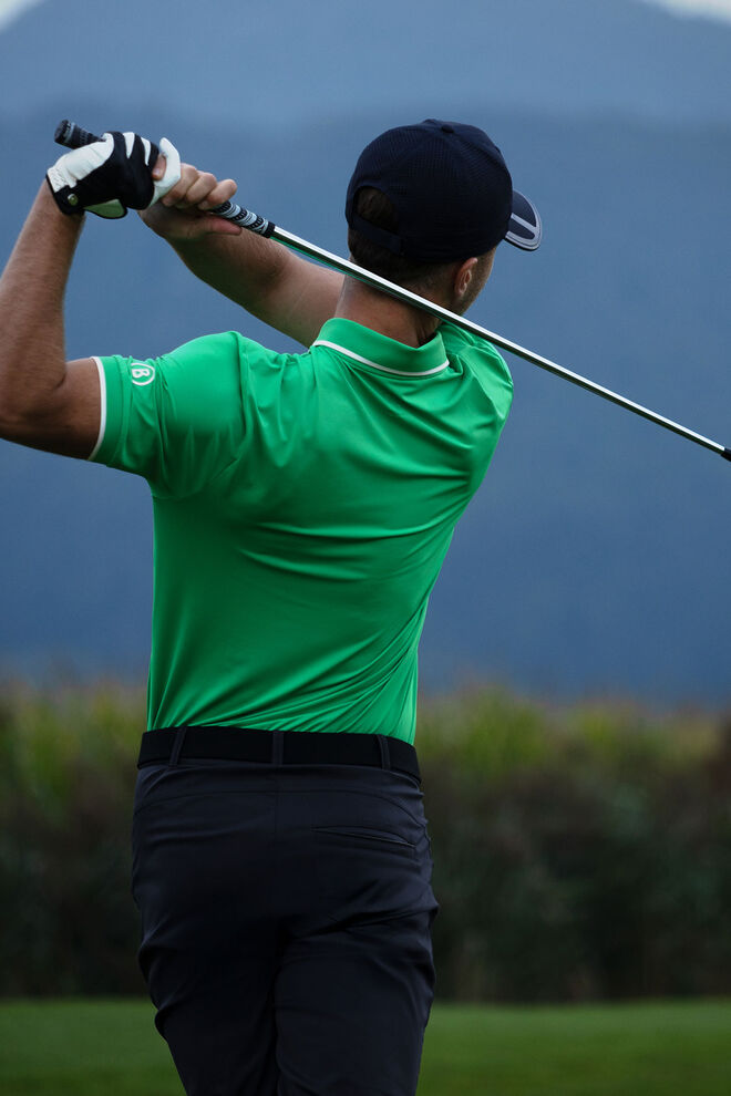 The male golfer is shown from the backside. He wears a bright green polo shirt and navy blue pants and tees off.
