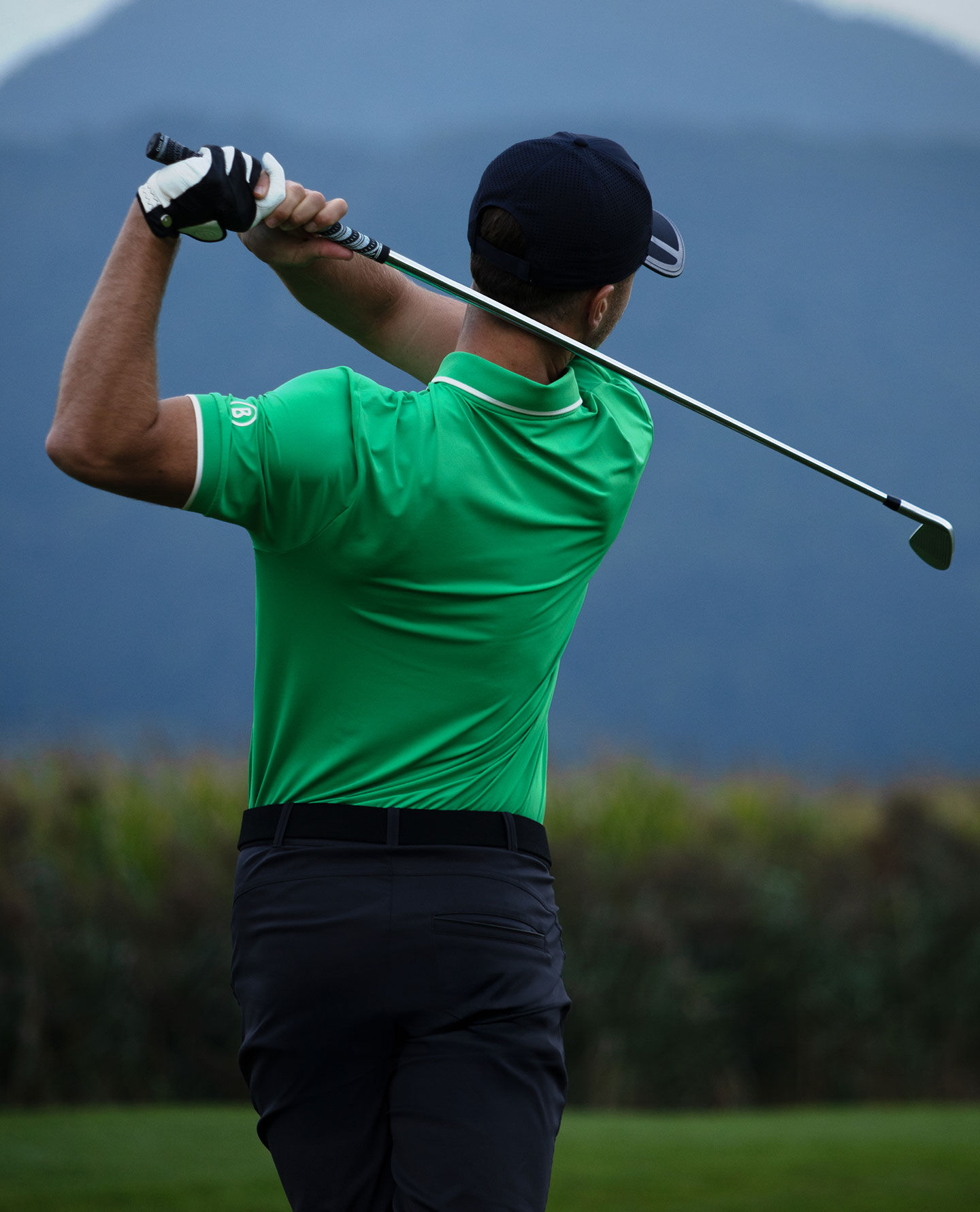 A golfer teeing off, photographed from behind. He is wearing a bright green polo shirt and dark trousers.