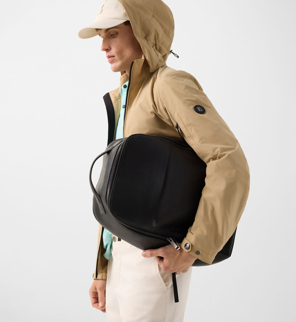 The male model wears a cyan blue polo shirt and a beige parka jacket. He carries a black leather backpack.