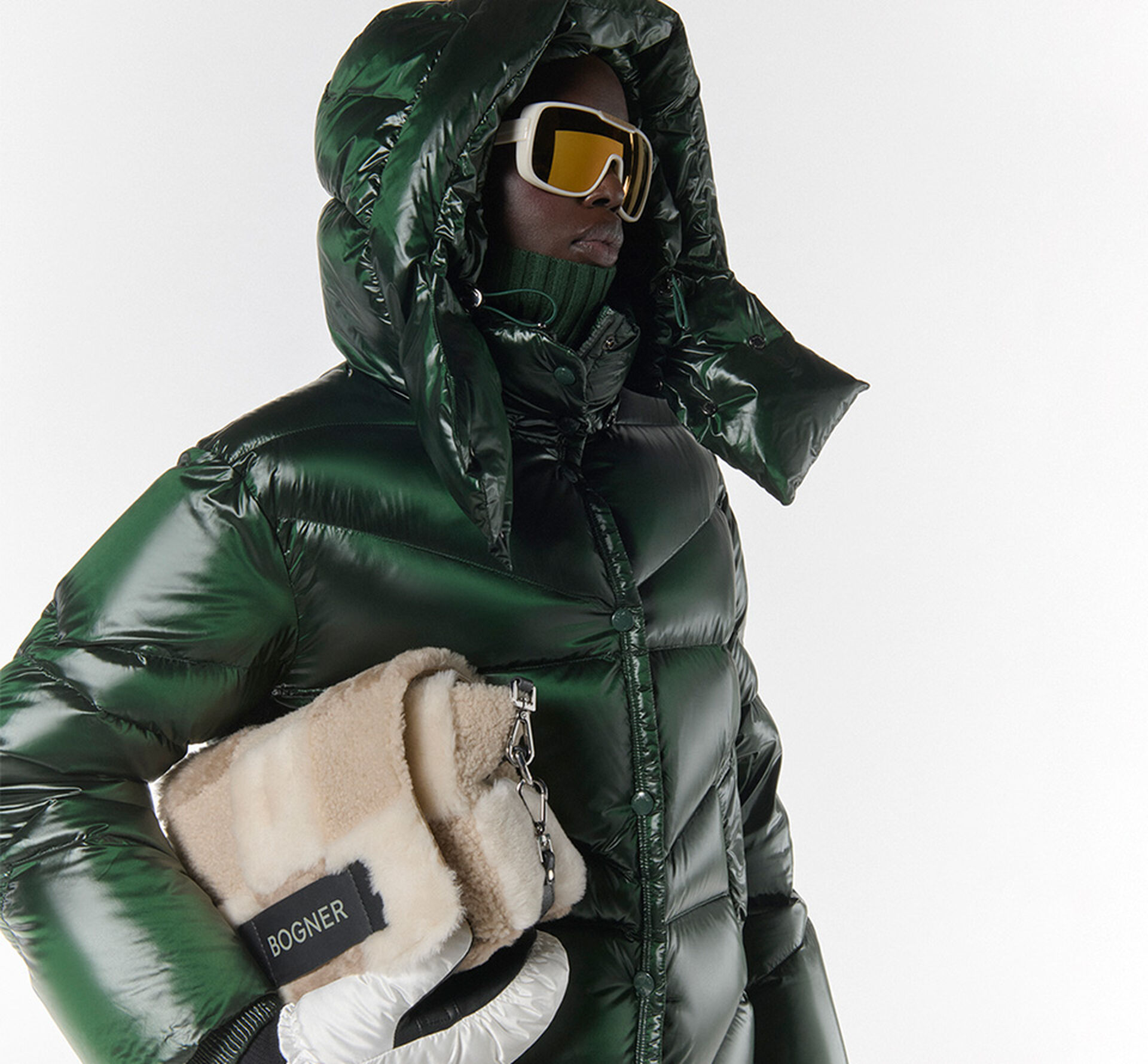 The female model wears a green down jacket and sunglasses. She holds a fur-trimmed bag in her arms.