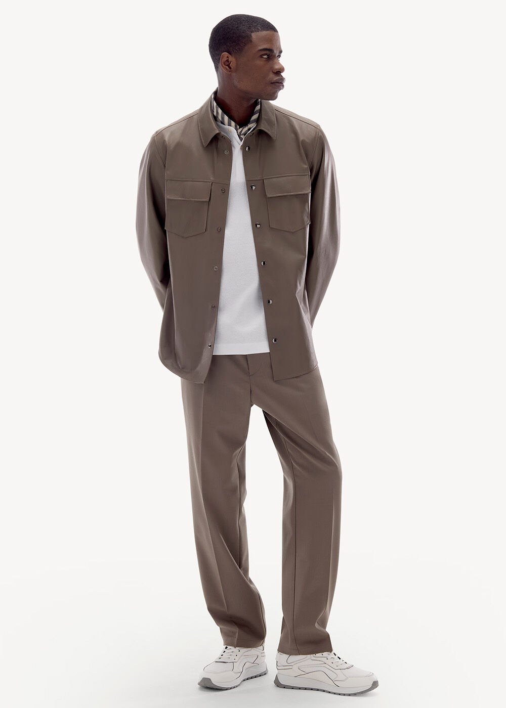 Ganzkörperaufnahme des Models mit weißem Polo, Leder-Overshirt und Sweatpants in Mud, dazu weiße Sneaker.