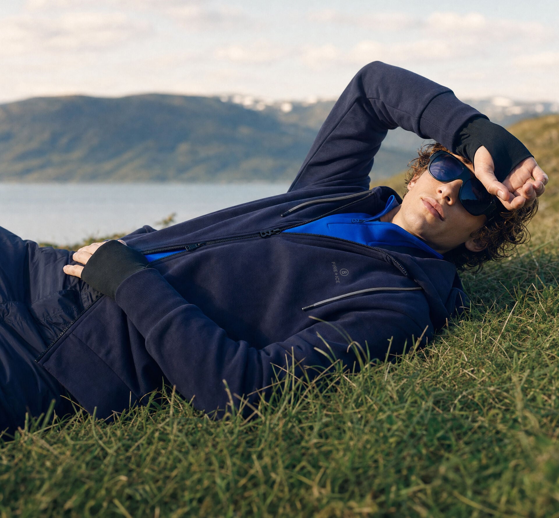 Das Herren Model liegt in einer Graslandschaft vor Bergkulisse. Er tr&auml;gt eine dunkelblaue Funktionsjacke mit royalblauem Shirt darunter.