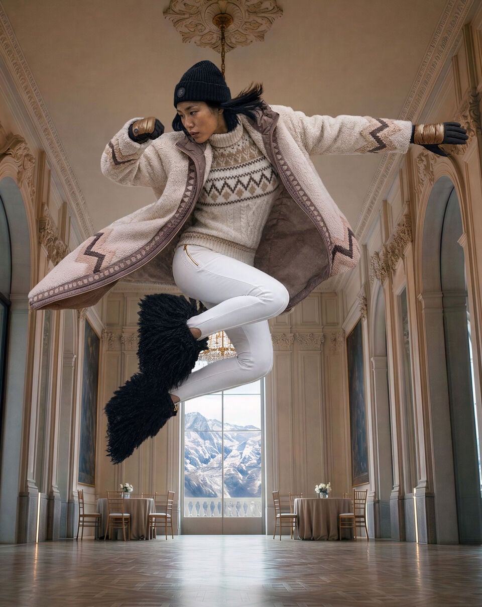 Das Model in dynamischer Pose tr&auml;gt einen gemusterten Mantel &uuml;ber einem Norwegerpullover zu wei&szlig;er Steghose und Fellstiefeln vor alpiner Bergkulisse.