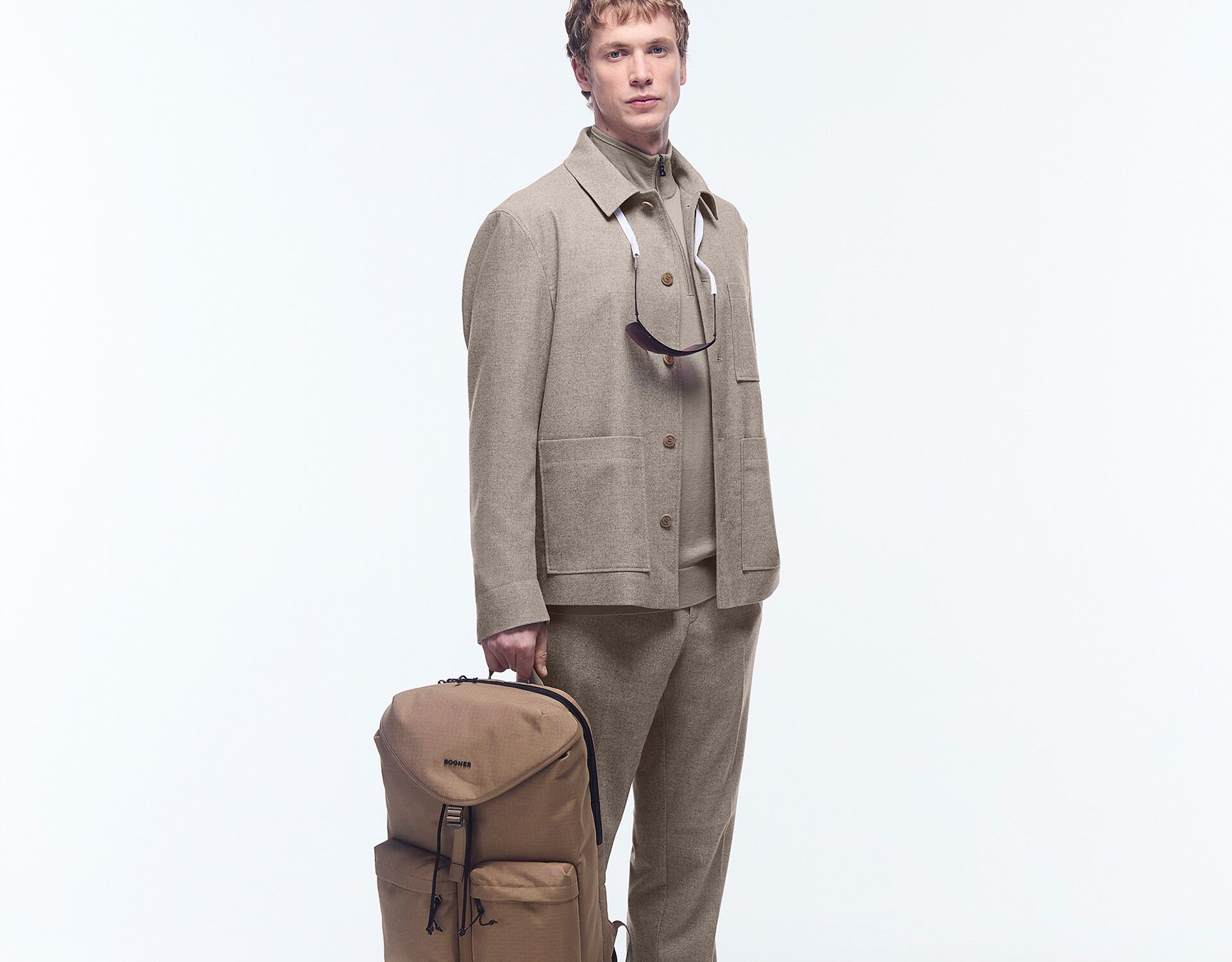The male model wears a beige outfit, a spacious BOGNER backpack and sunglasses around his neck.
