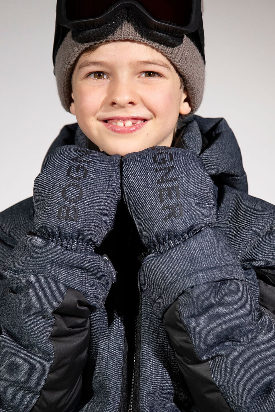 The boy is wearing a BOGNER ski jacket in performance denim, with matching gloves, a brown woolly hat and ski goggles.