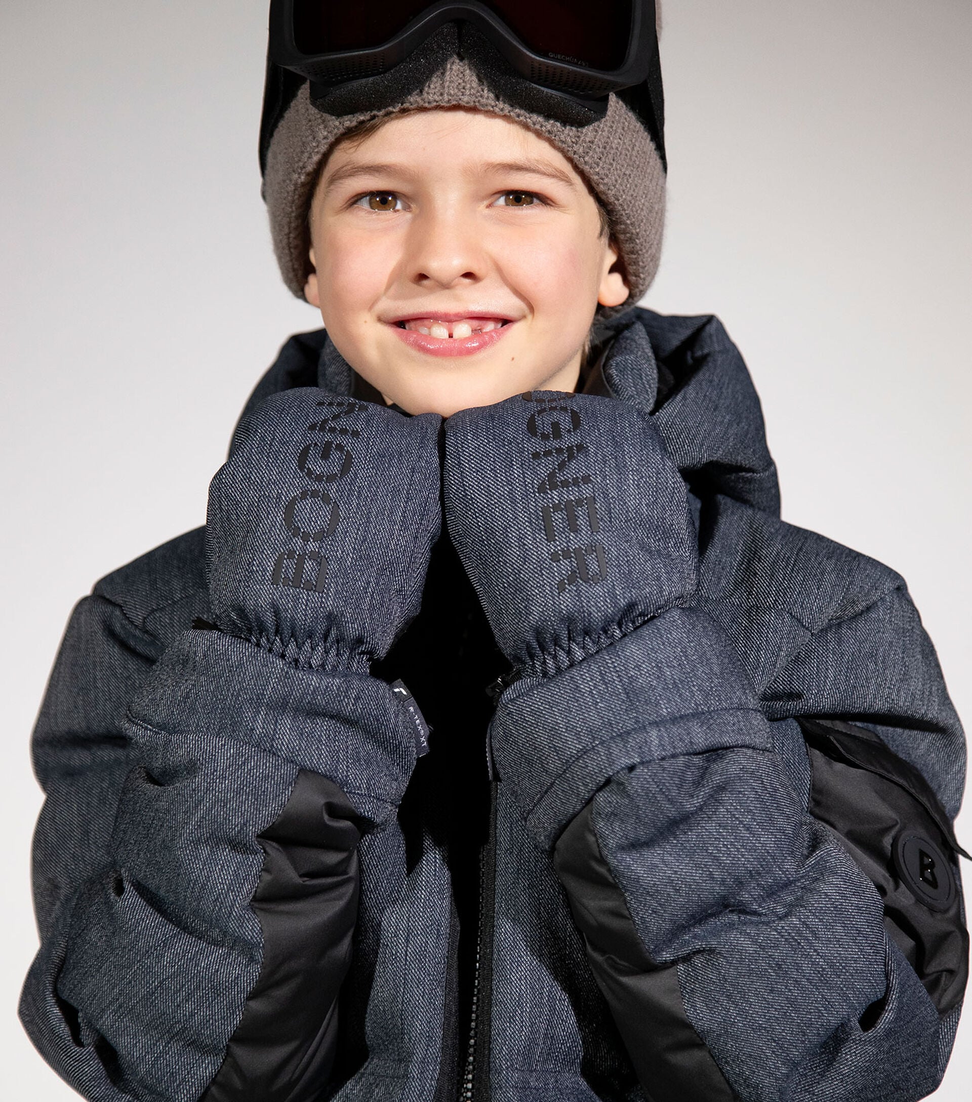 The boy is wearing a BOGNER ski jacket in performance denim, with matching gloves, a brown woolly hat and ski goggles.