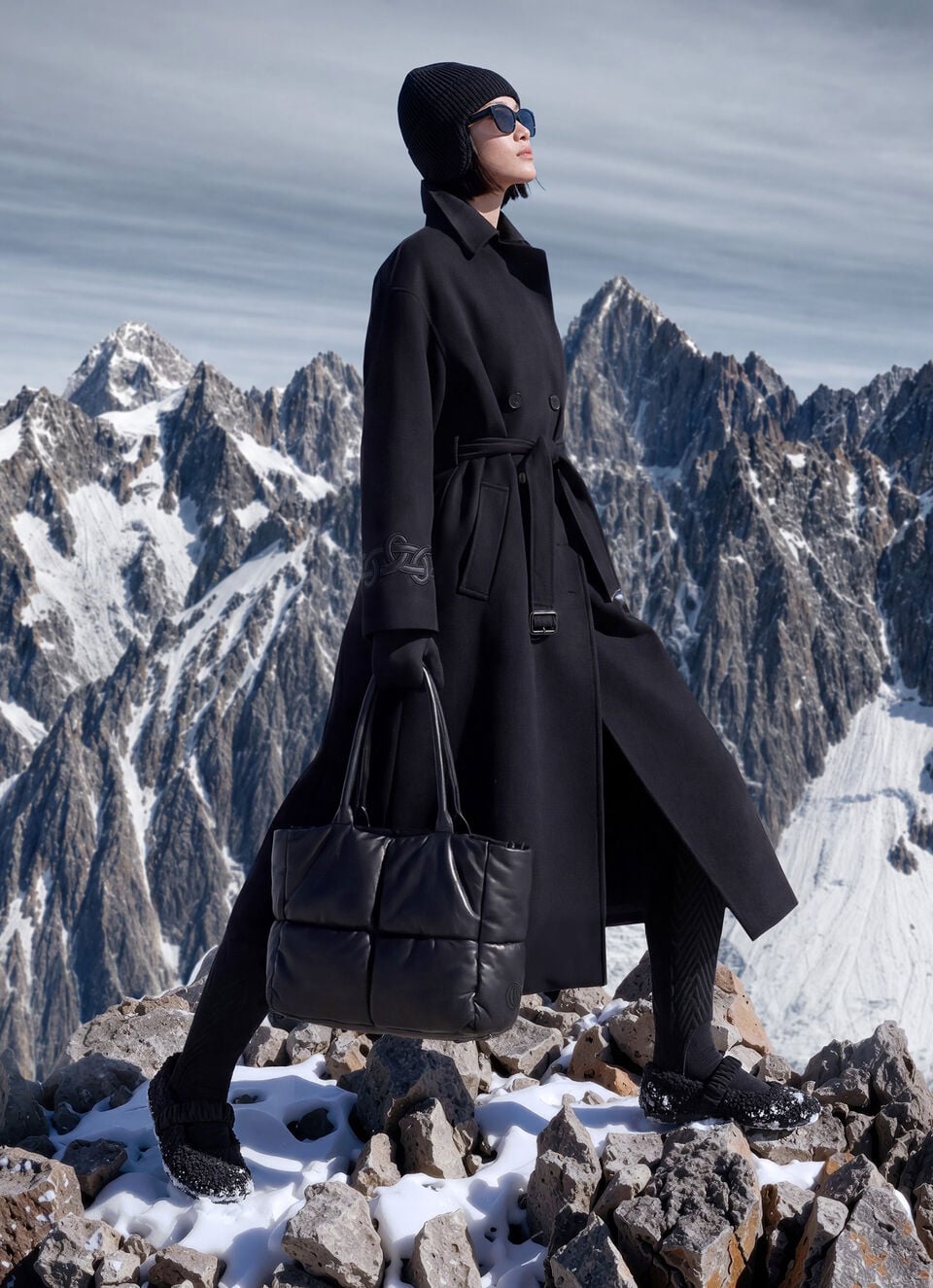 The female model with mountain scenery in the background is wearing a black coat with a wrap belt, and a knitted hat, sunglasses, a tote bag and teddy-fur ballerina flats to complete the look.