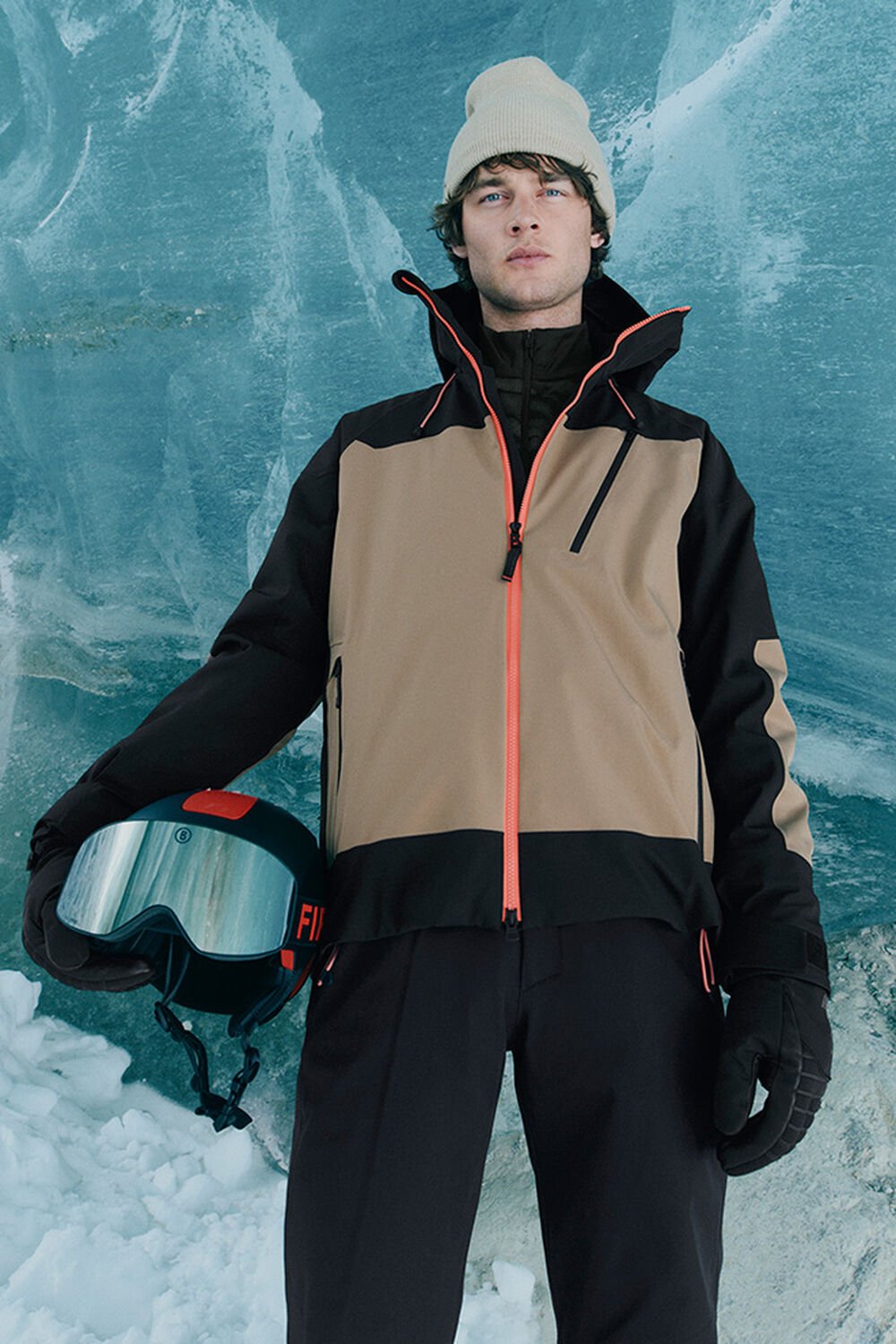 Hier trägt das Herren Model eine braun-schwarze Skijacke mit kontrastierenden Details. Im Hintergrund ist ein Gletscher.