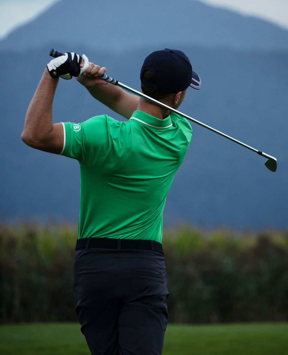 Seitliche Aufnahme des Herren Models beim Abschlag. Er tr&auml;gt ein Polo-Shirt in leuchtendem Gr&uuml;n und eine dunkle Hose.