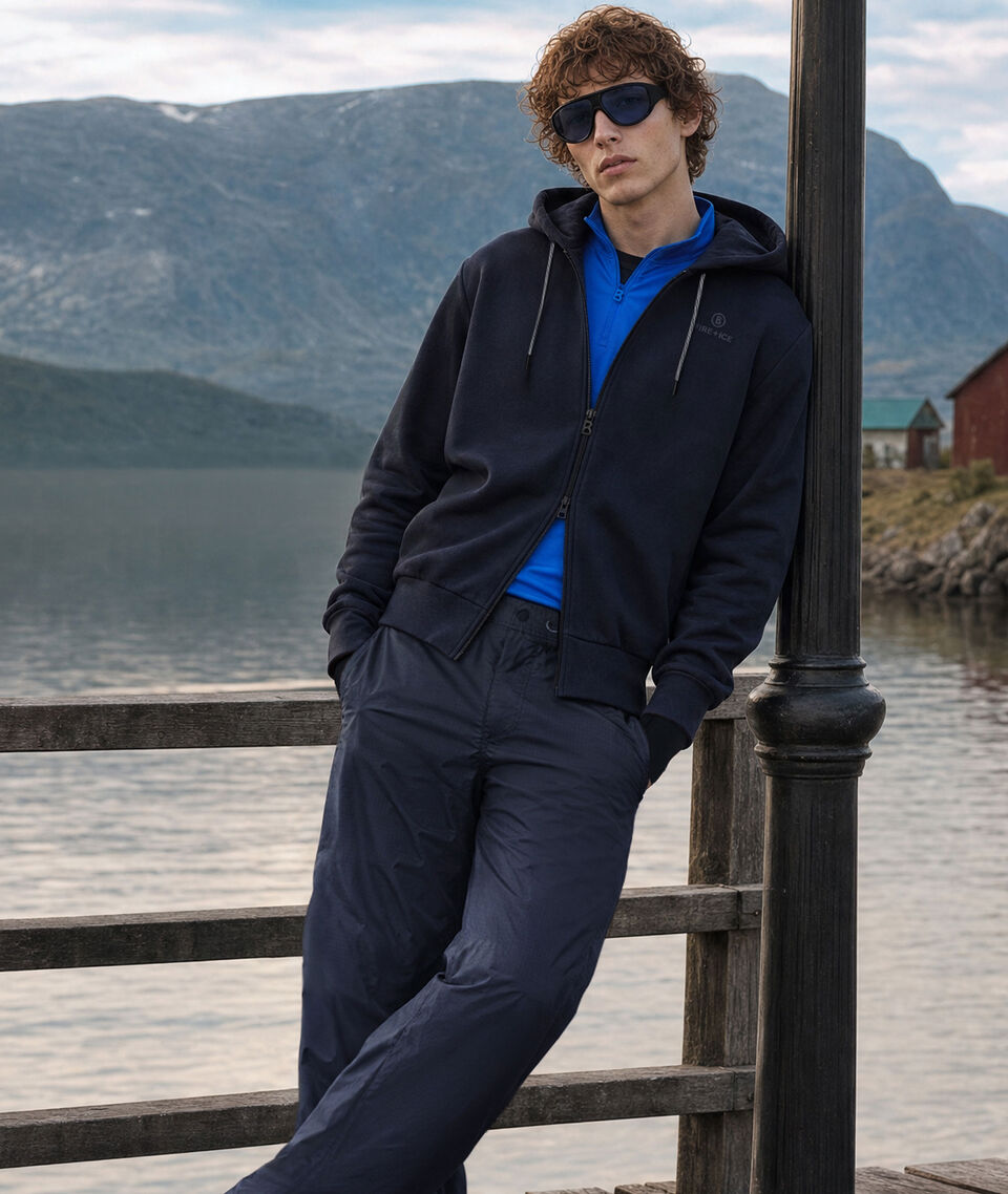 Das Herren Model steht vor einem Bergsee mit Gebirgskulisse. Er tr&auml;gt eine dunkelblaue Funktionsjacke und eine passende Hose, dazu First Layer Pascal darunter.