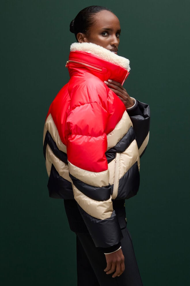 Le mannequin féminin porte une doudoune de ski aux larges motifs matelassés en W, mêlant noir, rouge et beige pour un style audacieux et chaud sur les pistes.