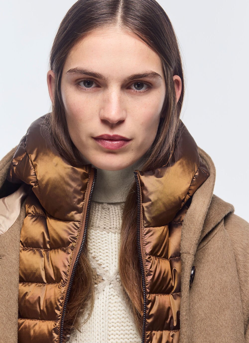 Close-up of a shiny down vest with a hooded coat over it and a grey turtleneck sweater underneath.