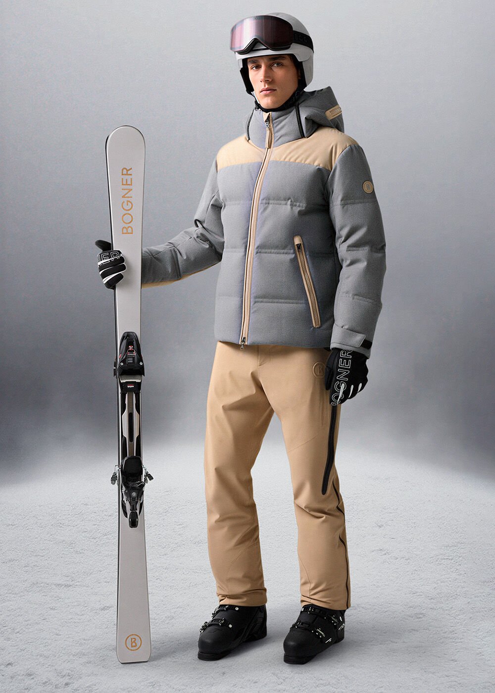 Das Herren-Model trägt eine grau-beigefarbene Skijacke, dazu eine beigefarbene Skihose und einen Skihelm.