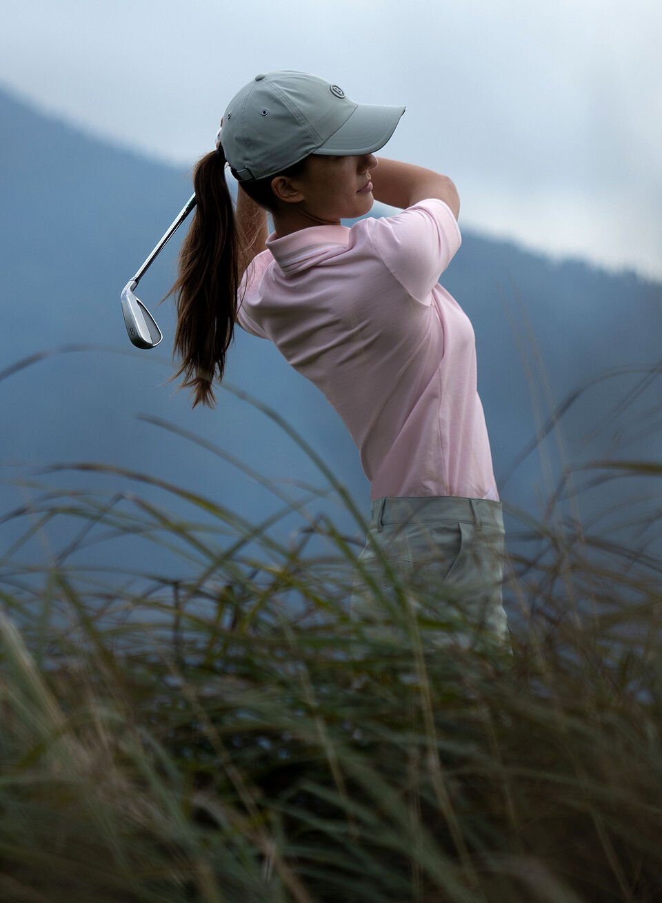 A side view half-body shot of a female golfer teeing off wearing a pink polo shirt. Mountain scenery behind.