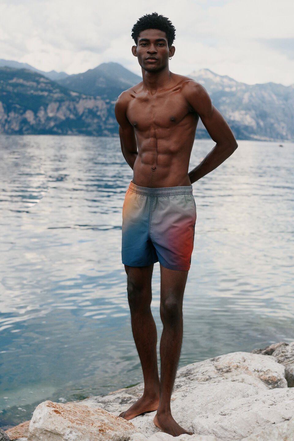 The male model is wearing colourful printed swimming trunks. There are mountains and a mountain lake in the background.