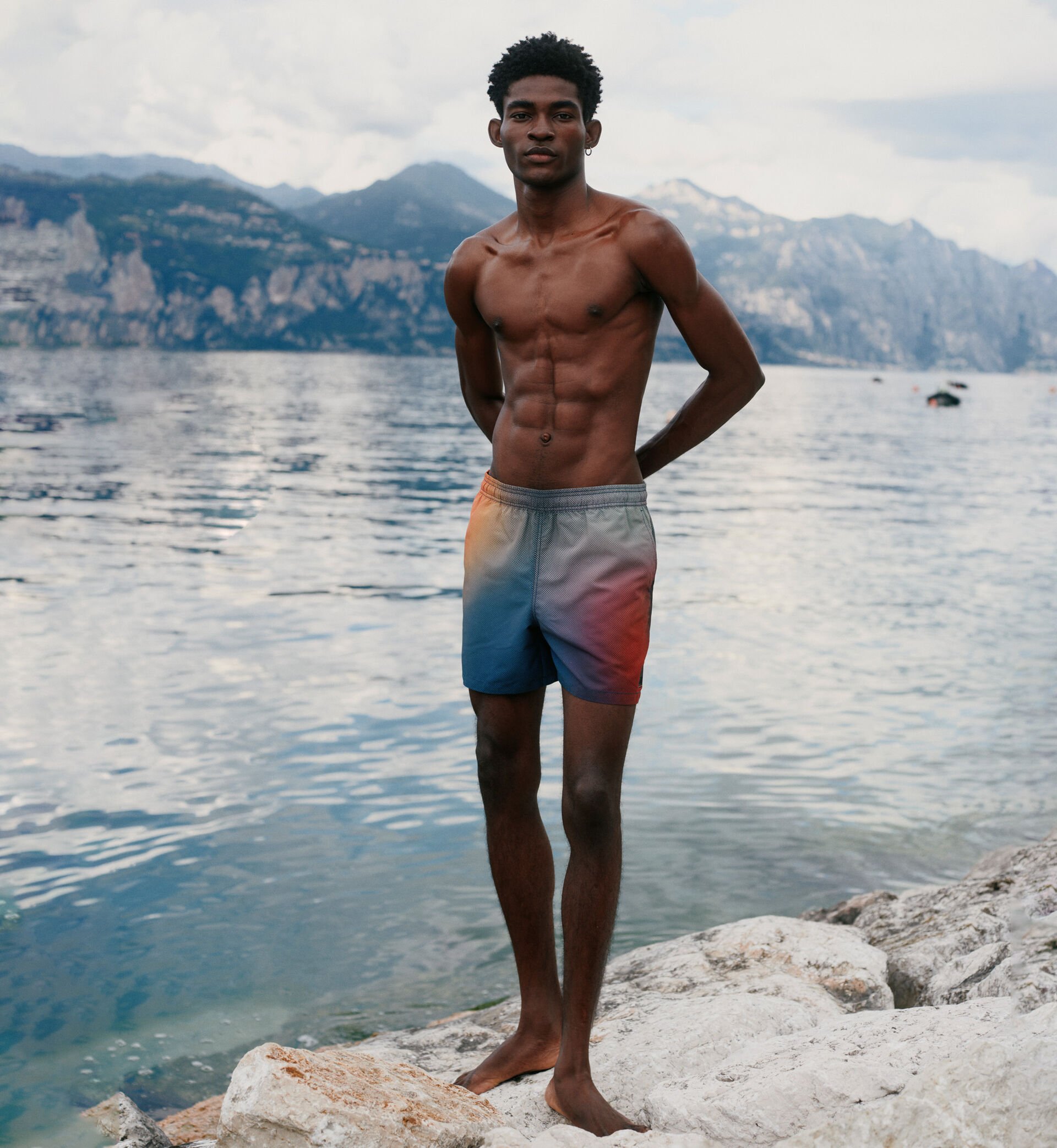 The male model is wearing colourful printed swimming trunks. There are mountains and a mountain lake in the background.