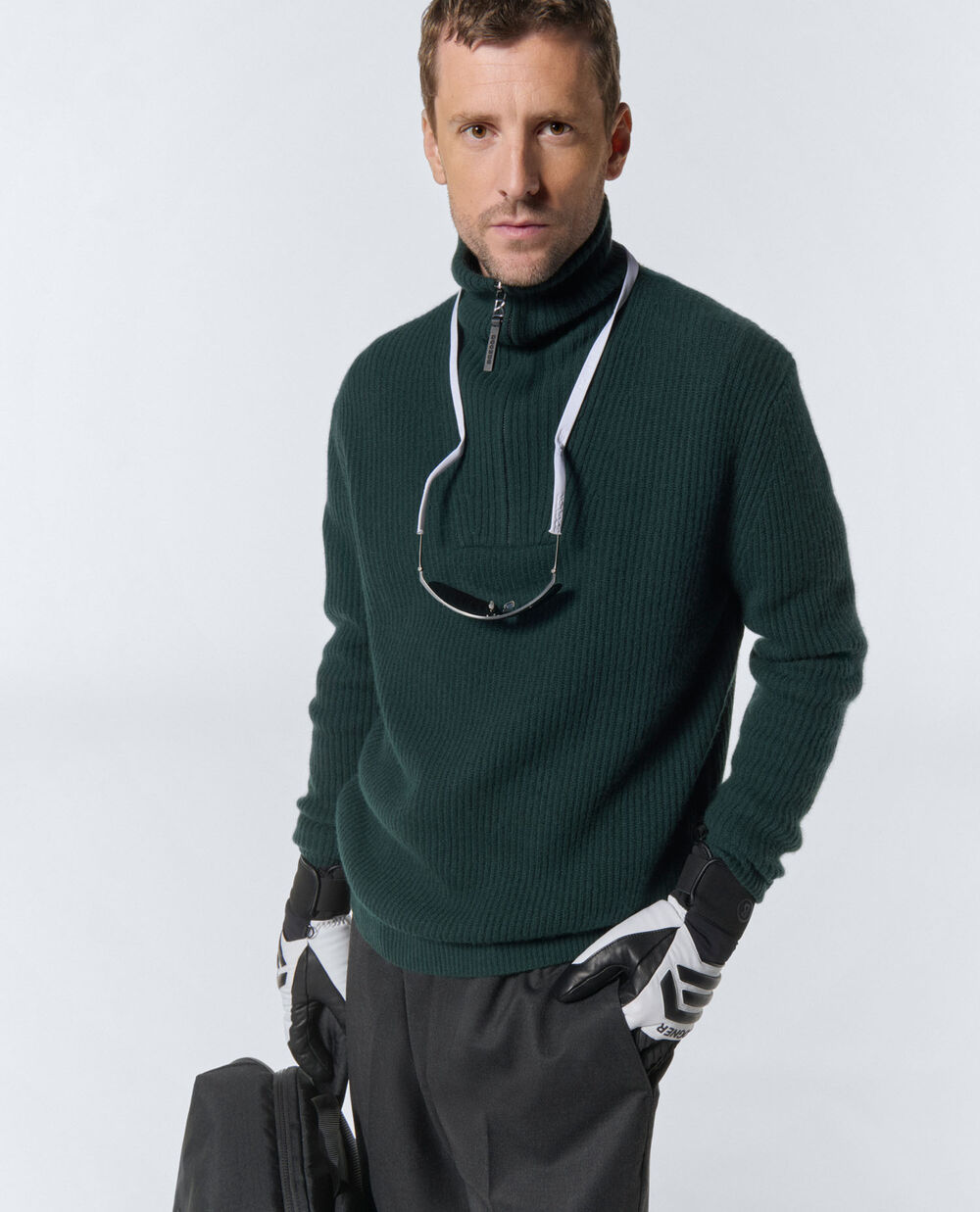 The model is wearing a dark green half-zip pullover with a stand-up collar, combined with dark grey pleat-front trousers in wool. 